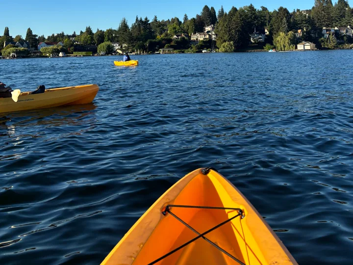 The bow of a yellow kayak on Union Bay, other kayakers and the wooded shoreline ahead
